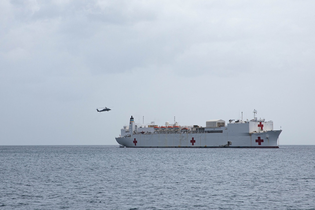 A white U.S. military hospital ship floats in the ocean while a military helicopter approaches it
