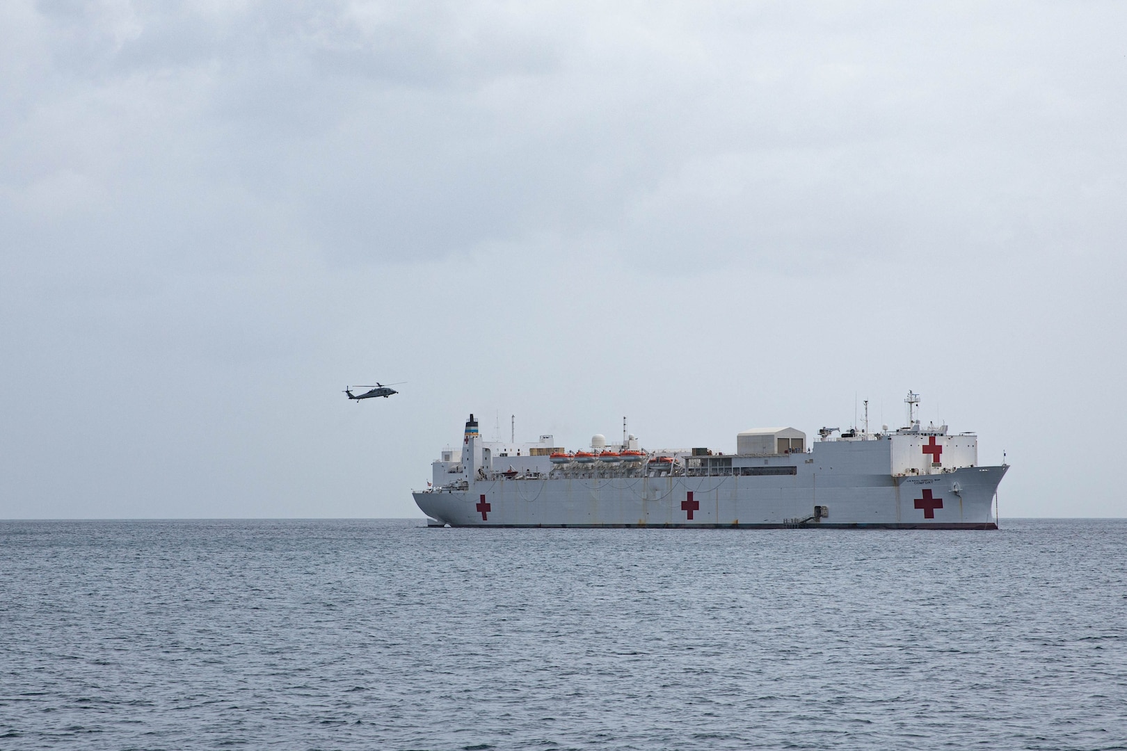 A white U.S. military hospital ship floats in the ocean while a military helicopter approaches it