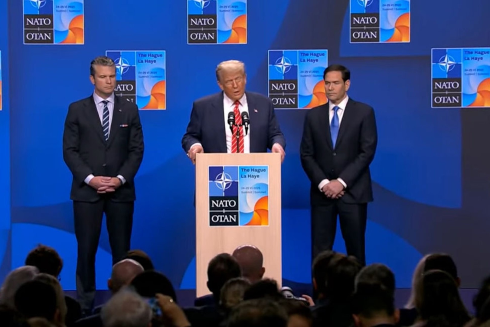 A man in a suit and tie speaks in front of a lectern, with two men in suits standing on either side of him.