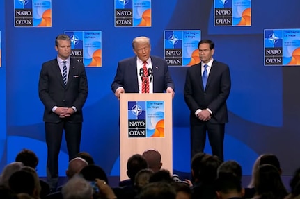 A man in a suit and tie speaks in front of a lectern, with two men in suits standing on either side of him.