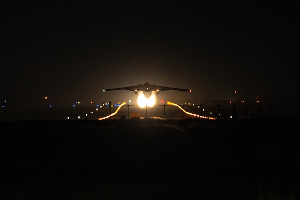 A B-2 bomber aircraft lifts off from a dark runway, trailing two fire-like contrails with lights in the background.