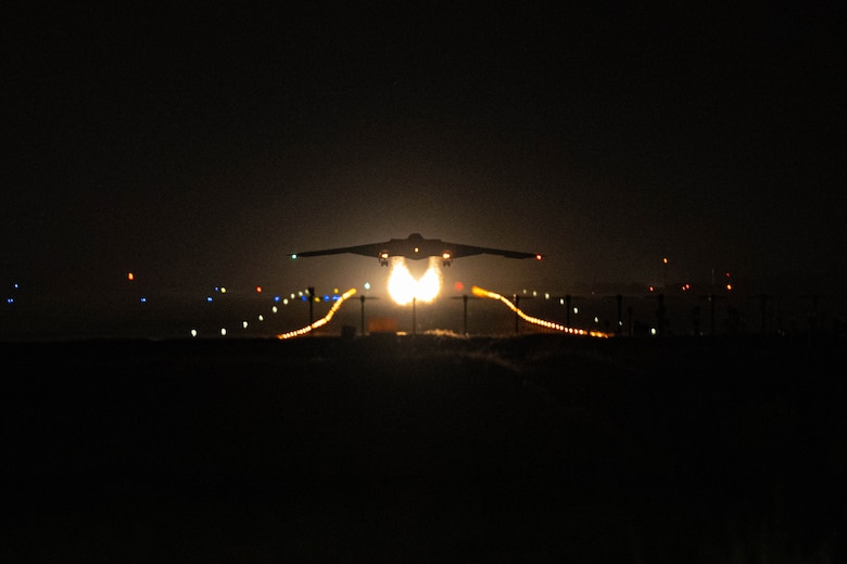 A B-2 bomber aircraft lifts off from a dark runway, trailing two fire-like contrails with lights in the background.