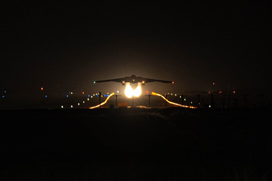 A B-2 bomber aircraft lifts off from a dark runway, trailing two fire-like contrails with lights in the background.