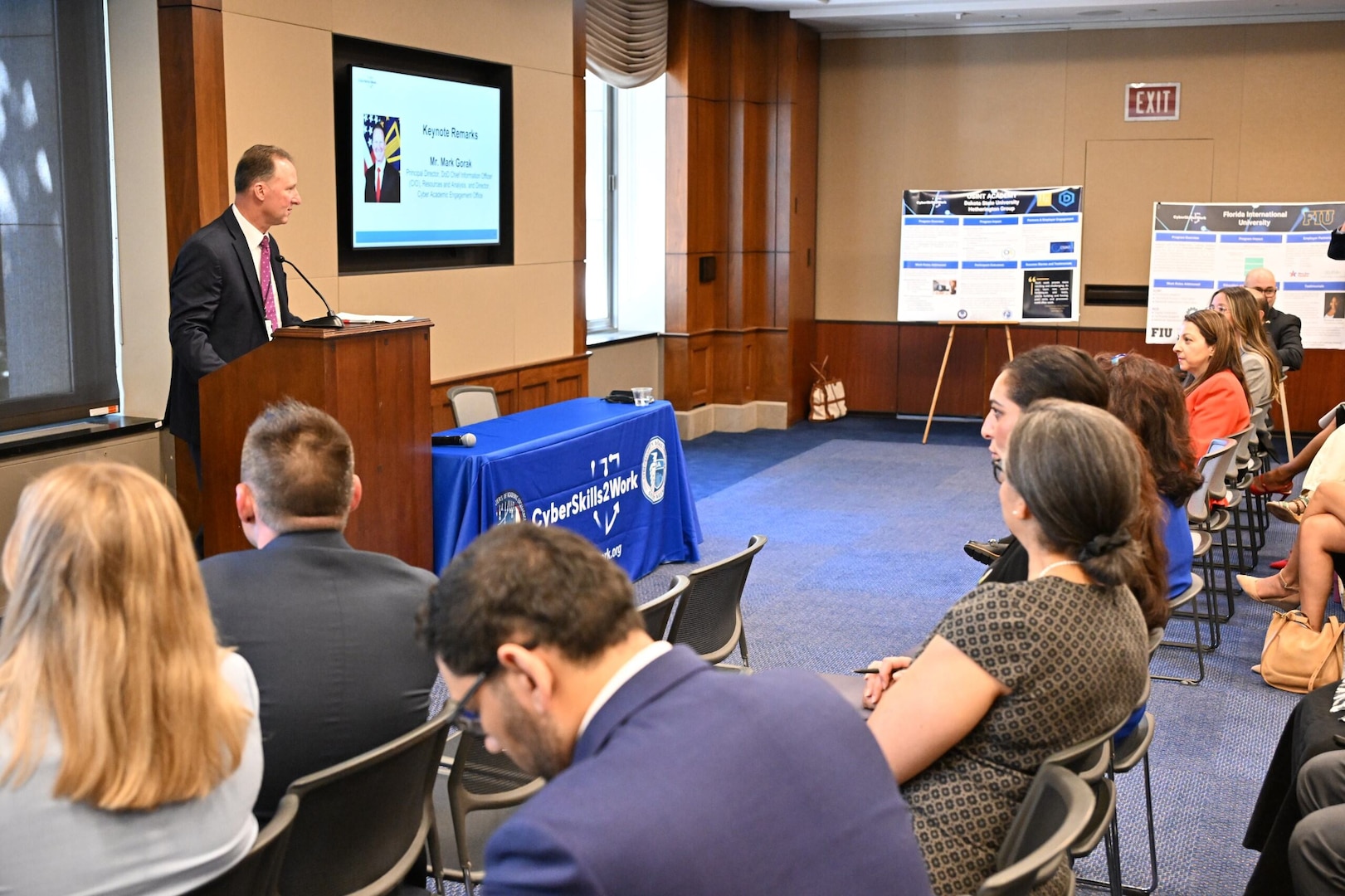 A man in business attire is standing behind a podium in a room, speaking to people seated in chairs in front of him.
