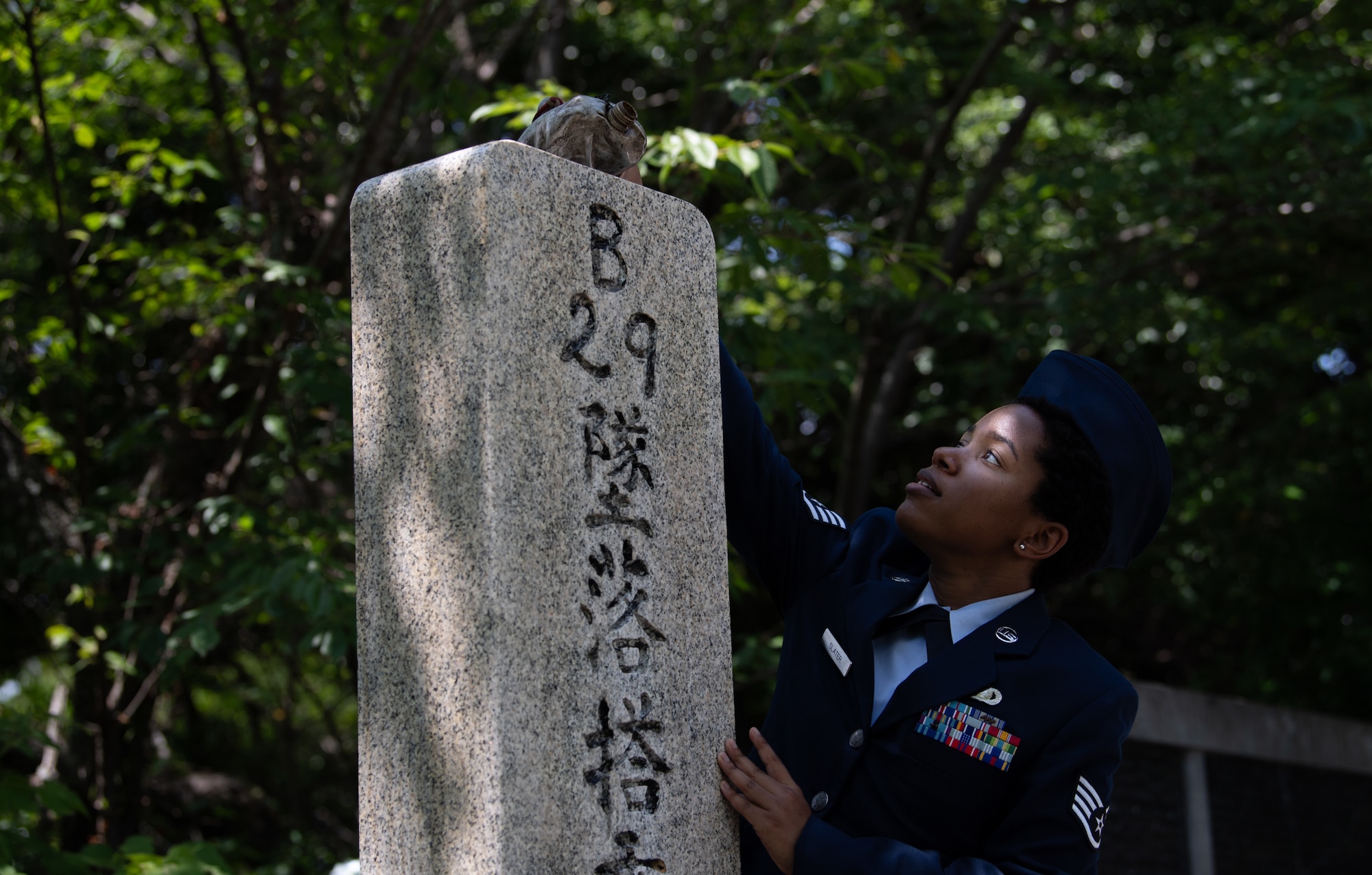 An Airman dressed in their blue dress uniform and flight cap reaches pours whiskey out of a canteen over a stone monument.