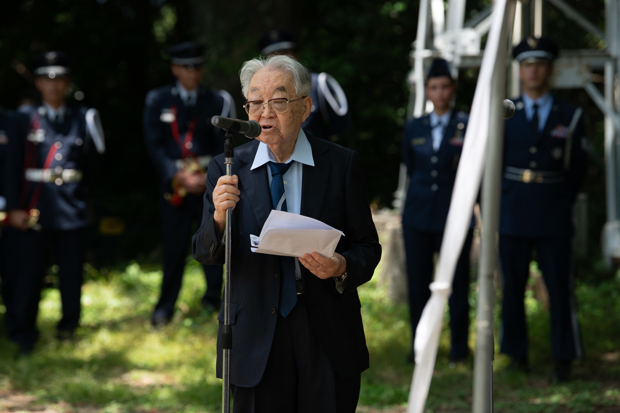 An elderly person with white hair grabs a microphone stand as he reads from a paper in his hand.