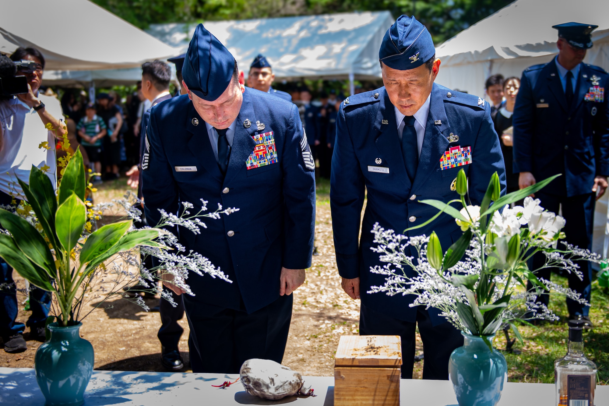 Two Airmen dressed in their blue dress uniform bow toward a monument decorated in flower vases and flowers.
