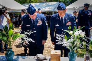 Two Airmen dressed in their blue dress uniform bow toward a monument decorated in flower vases and flowers.