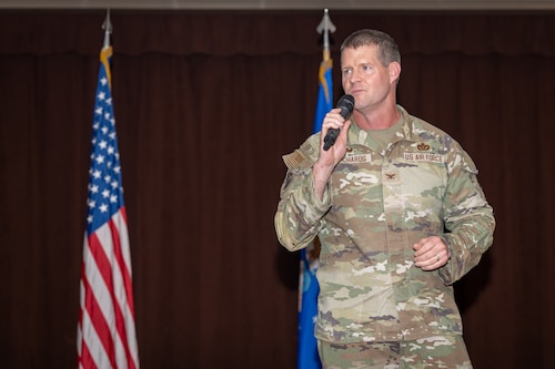 A man wearing camouflaged military uniform speaks into a microphone in front of the U.S. and Air Force flags