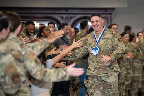 A smiling camouflaged uniformed military member, wearing a master sgt. chevron around his neck, high fives other military members