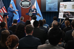 A man wearing business attire stands next to another wearing a formal military uniform in front of a group of people. Both stand at podiums, looking at a poster displayed on an easel with pictures on it. There is a sign between them that reads, “The Pentagon.”