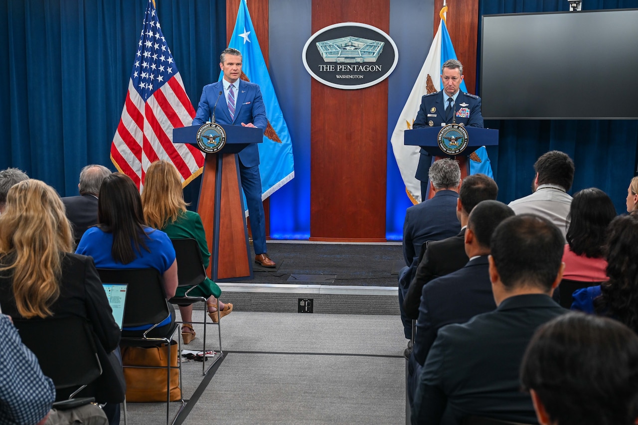 Two men stand at podiums in front of a group of people. The man on the left is wearing business attire, and the man on the right is wearing a formal military uniform. There is a sign between them that reads, “The Pentagon,” and an American flag is displayed behind them to the left.