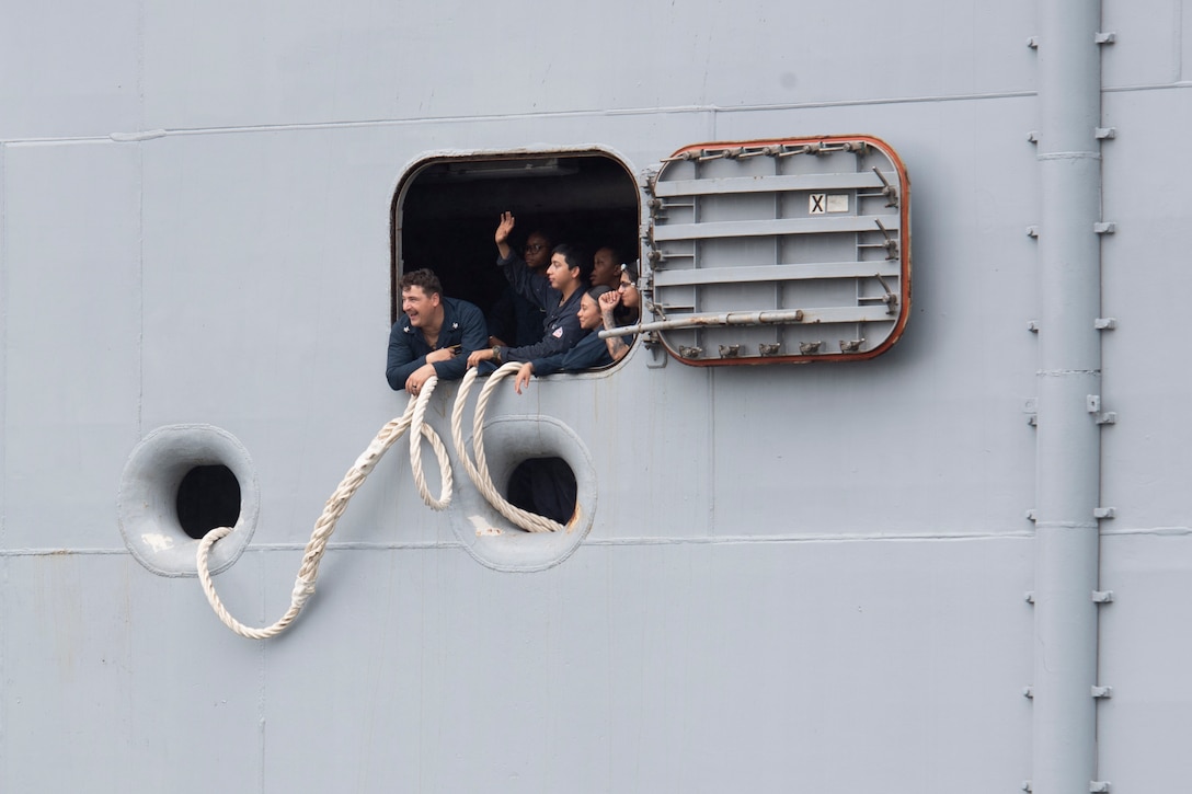 Sailors wave through an open hatch on a grey ship.