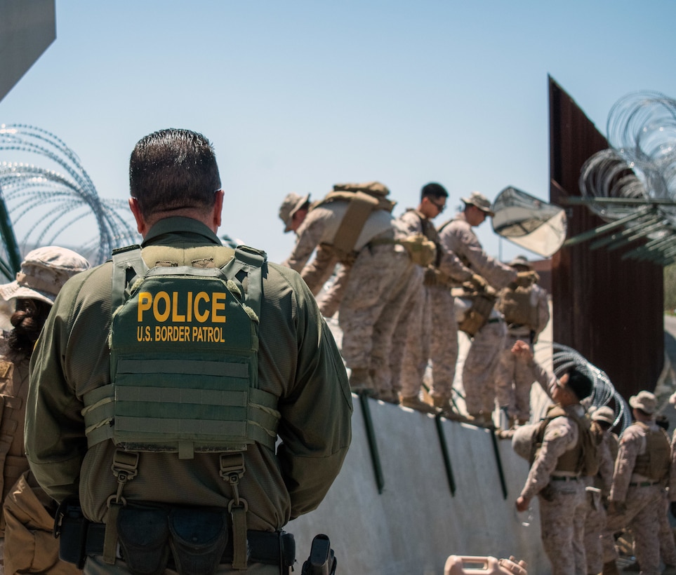 A Border Patrol agent watches a group of Marines install barbed wire on a security wall on a sunny day.