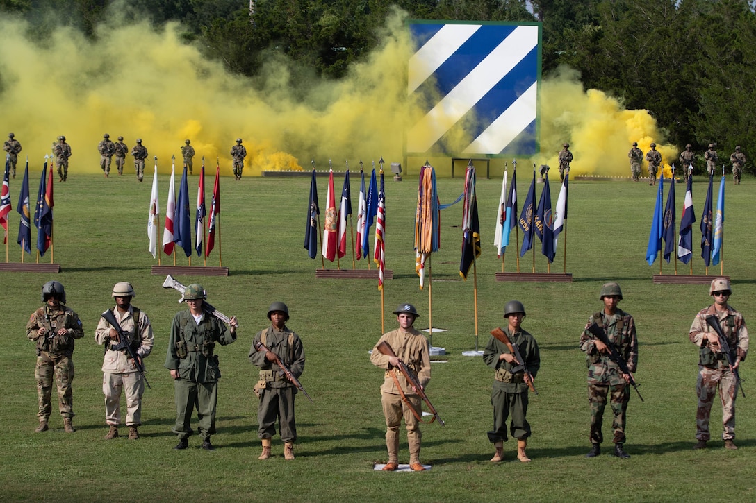 Soldiers in historic uniforms stand in front of standing flags on a field with yellow smoke behind.
