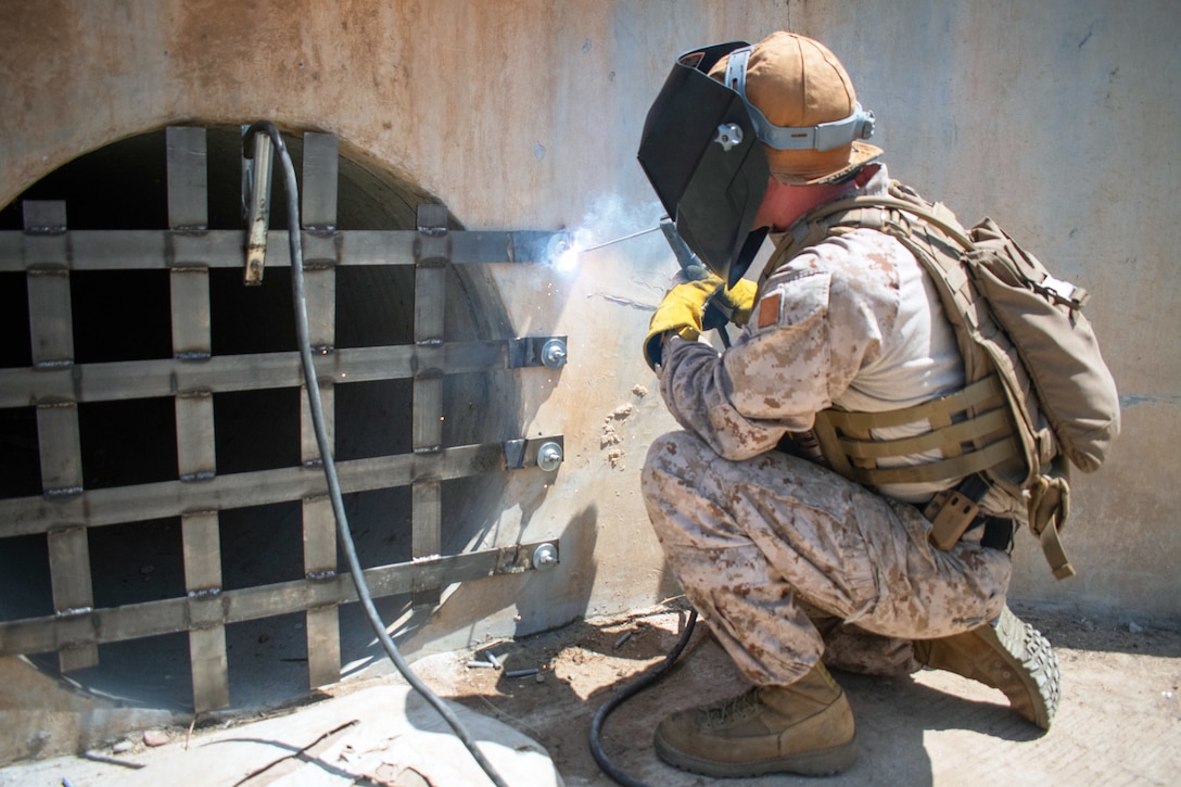 A Marine wearing a protective visor welds a metal gate to the opening of a concrete tunnel outdoors.