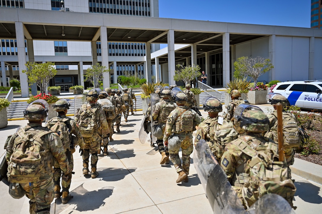 Soldiers in combat gear and carrying shields walk on pavement toward an office building with a police vehicle parked nearby.