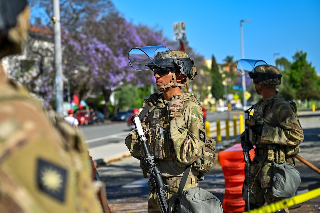 Three soldiers in combat gear stand near a street on a sunny day, with an orange security barrier and yellow tape also visible.