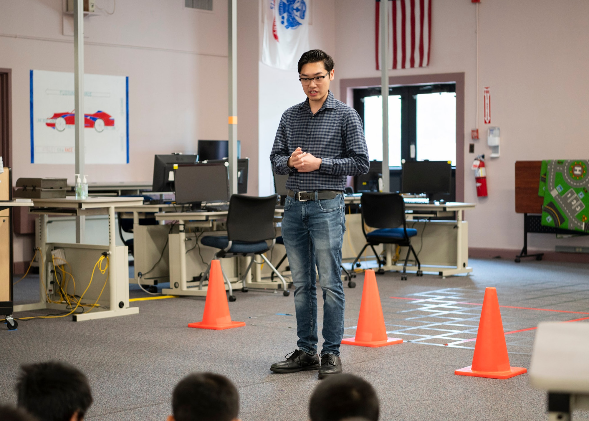 A man with glasses stands near computer desks while speaking to an audience.