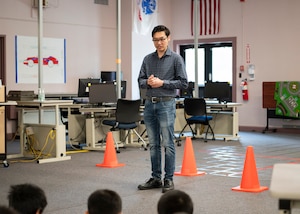 A man with glasses stands near computer desks while speaking to an audience.