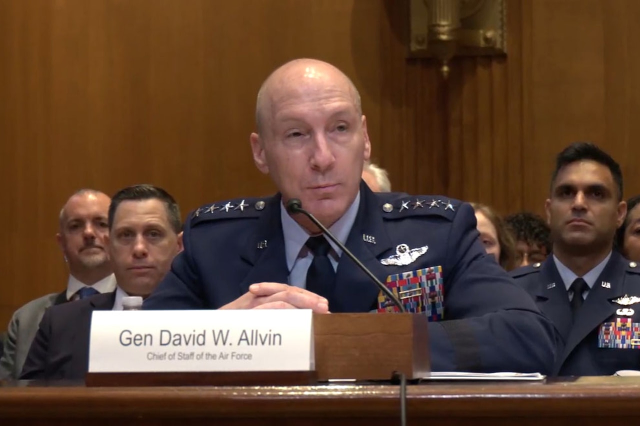 A man wearing a military dress uniform sits at a table while speaking into a microphone during a hearing. Several people in military dress uniforms and business attire sit behind him.