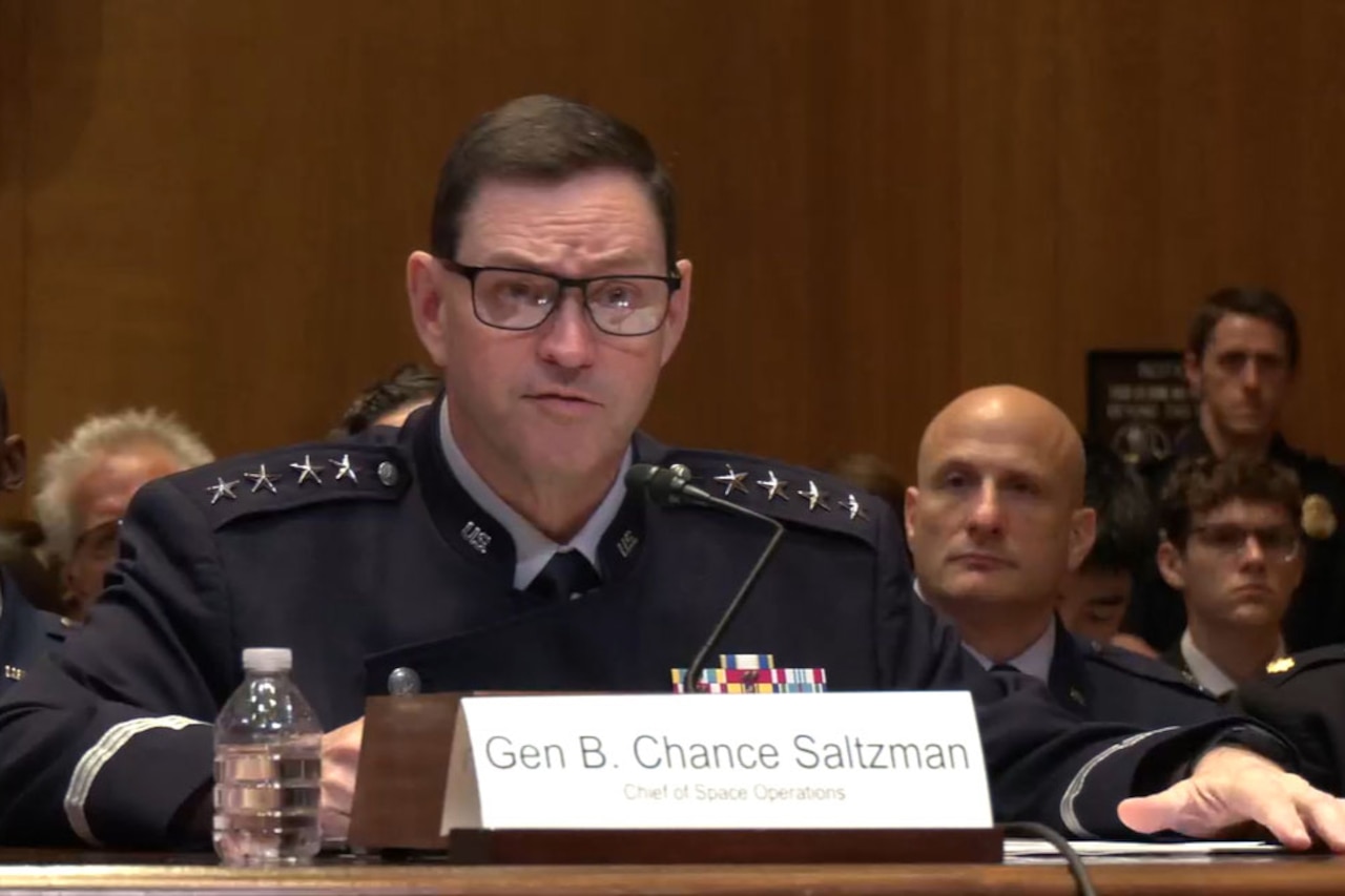 A man wearing a military dress uniform sits at a table while speaking into a microphone during a hearing. Several people in military dress uniforms and business attire sit behind him.
