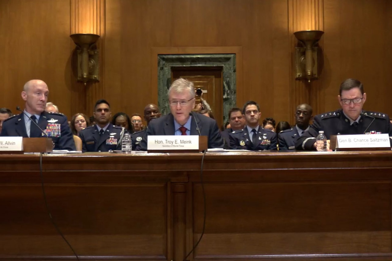Two men in military dress uniforms sit at a table while a man in business attire speaks into a microphone during a hearing. Several people in military dress uniforms and business attire sit behind him.