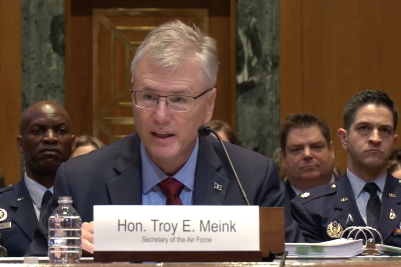 A man wearing business attire sits at a table while speaking into a microphone during a hearing. Several people in military dress uniforms sit behind him.