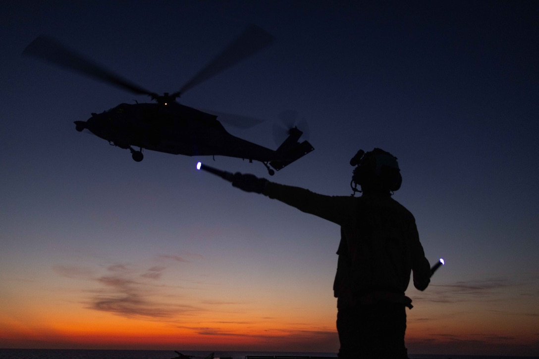 A sailor in silhouette holds two signal wands with light at the tips, one straight ahead, as a helicopter takes off from the deck of a ship.