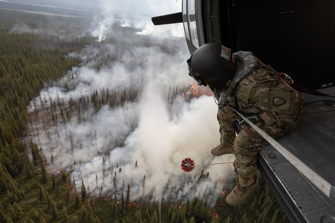 A soldier sits on the edge of a helicopter in flight over a forest fire and smoke.