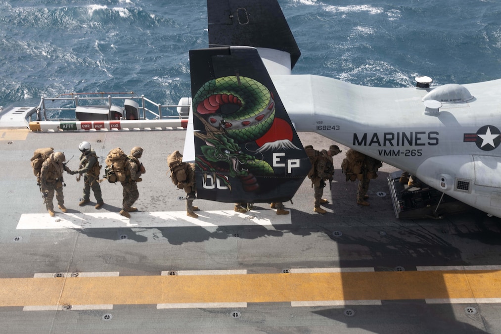 Marines with backpacks line up wand walk into an aircraft on a ship, as seen from above.
