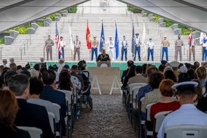 A uniformed man stand at a podium in front of a large crowd, with 12 soldiers lined up behind him holding flags.