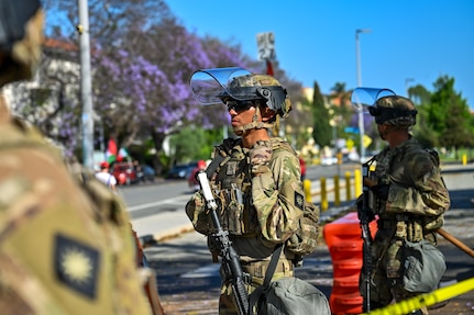 Three soldiers in combat gear stand near a street on a sunny day, with an orange security barrier and yellow tape also visible.