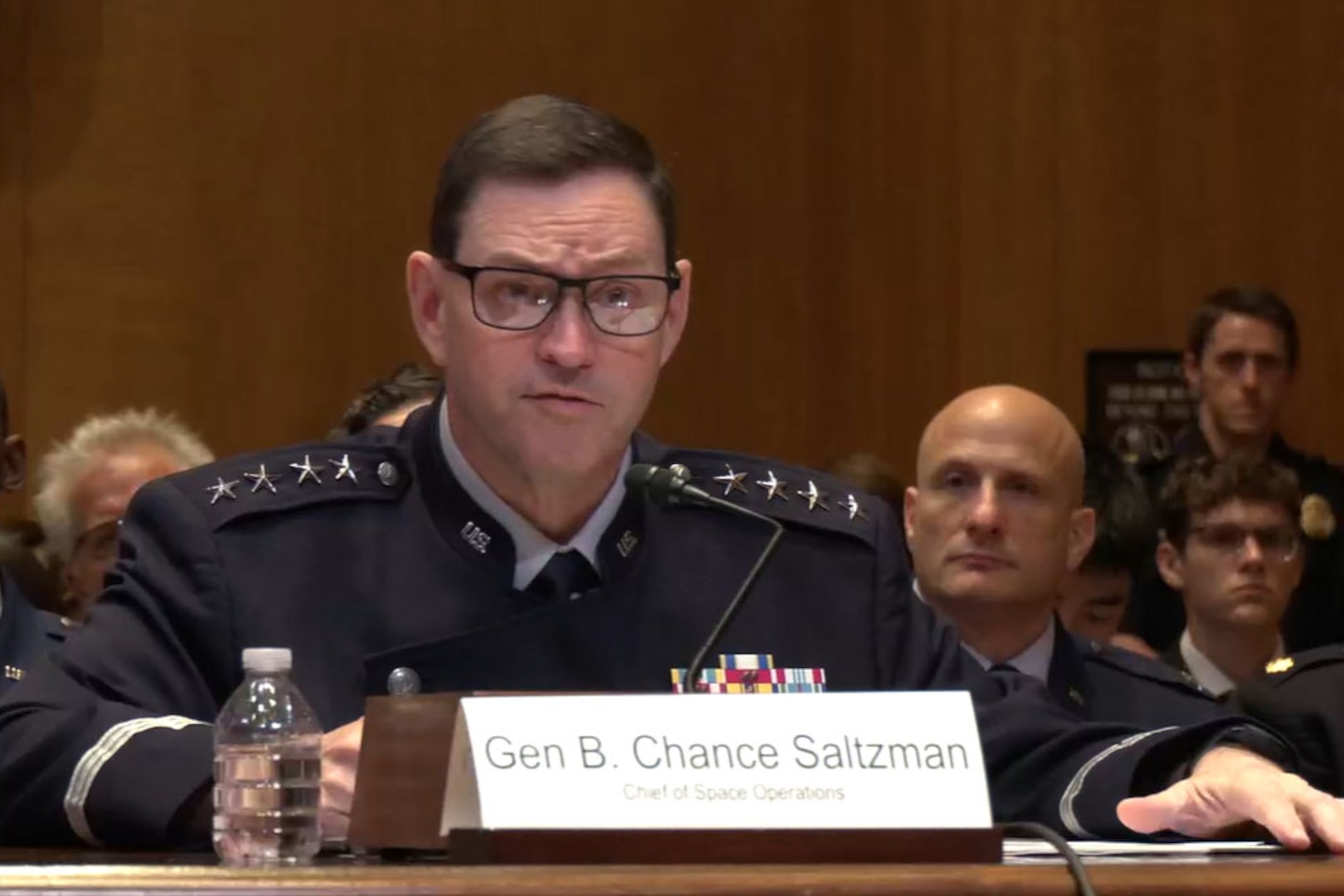 A man wearing a military dress uniform sits at a table while speaking into a microphone during a hearing. Several people in military dress uniforms and business attire sit behind him.