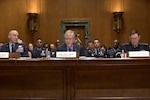Two men in military dress uniforms sit at a table while a man in business attire speaks into a microphone during a hearing. Several people in military dress uniforms and business attire sit behind him.