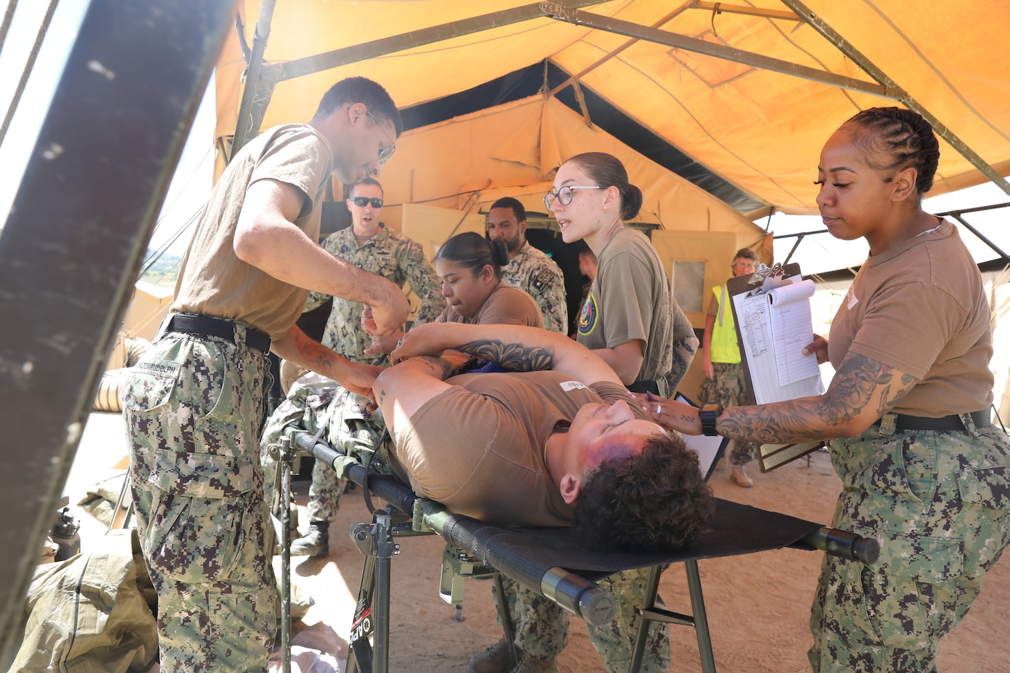 CS3 Kahlil Hamiltonrudolph (left) Culinary Specialist 3rd Class Estephania Stokes (center), Hospital Corpsman 3rd Class Jazzelle Davis, Hospital Corpsman 2nd Class Alexis Potter (right), Hospital Corpsman 1st Class Joseph Armella (back left), and Hospital Corpsman 3rd Class Denneson Wilson (back right) from Expeditionary Medical Facility Juliet (EMF-J), treat Equipment Operator Constructionman Jayden Hernandez (center) as he portrays a simulated patient during EMF training at Naval Expeditionary Medicine Warfighter Development Center (NEMWDC) May 3-16, 2025.  (U.S. Navy photo by Petty Officer 2nd Class Moira Esquivel)