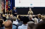 Man in blue suit standing to the left and woman in military uniform standing with a microphone to the right, speaking to an audience.