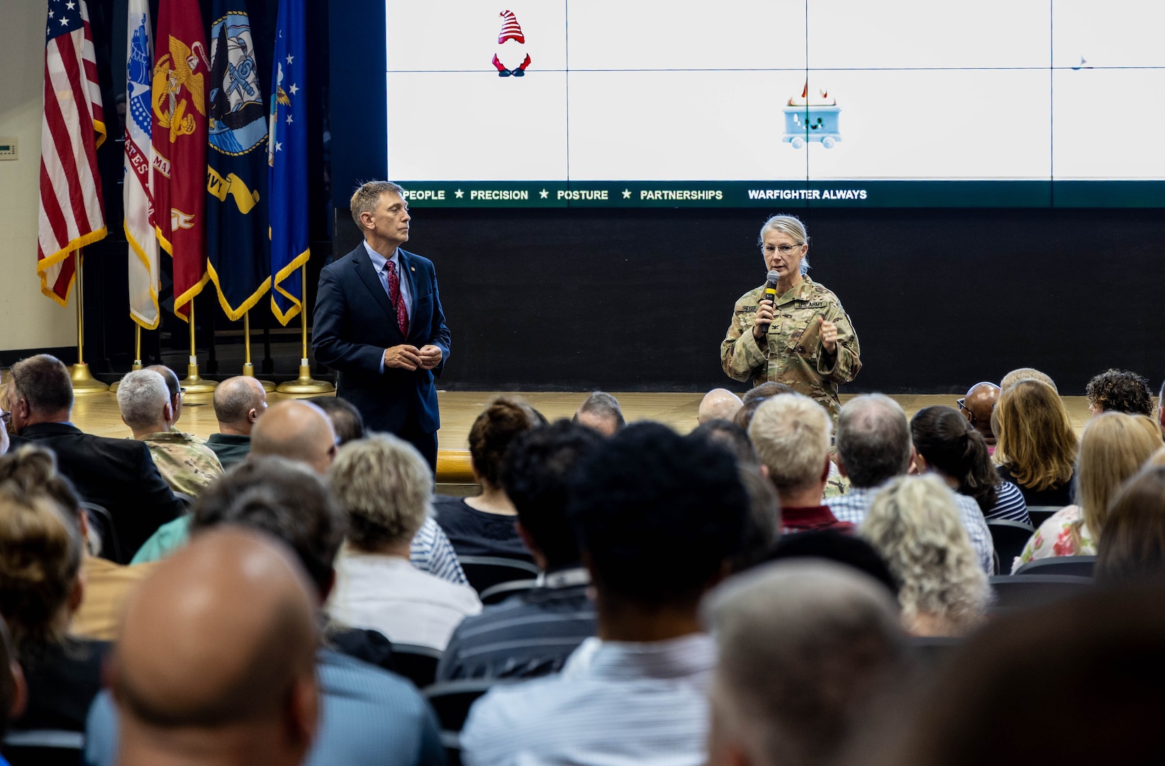 Man in blue suit standing to the left and woman in military uniform standing with a microphone to the right, speaking to an audience.