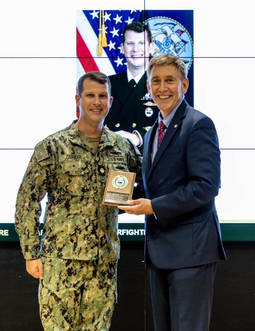 Man in military uniform on left and man in blue suit holding a plaque on right pose for photo, smiling. Headshot photo with flag in background.