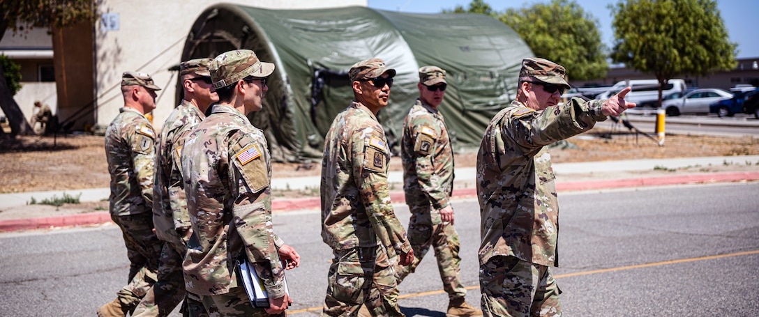 Six soldiers walk along a paved road on a sunny day with a green canopy and parked vehicles in the background.
