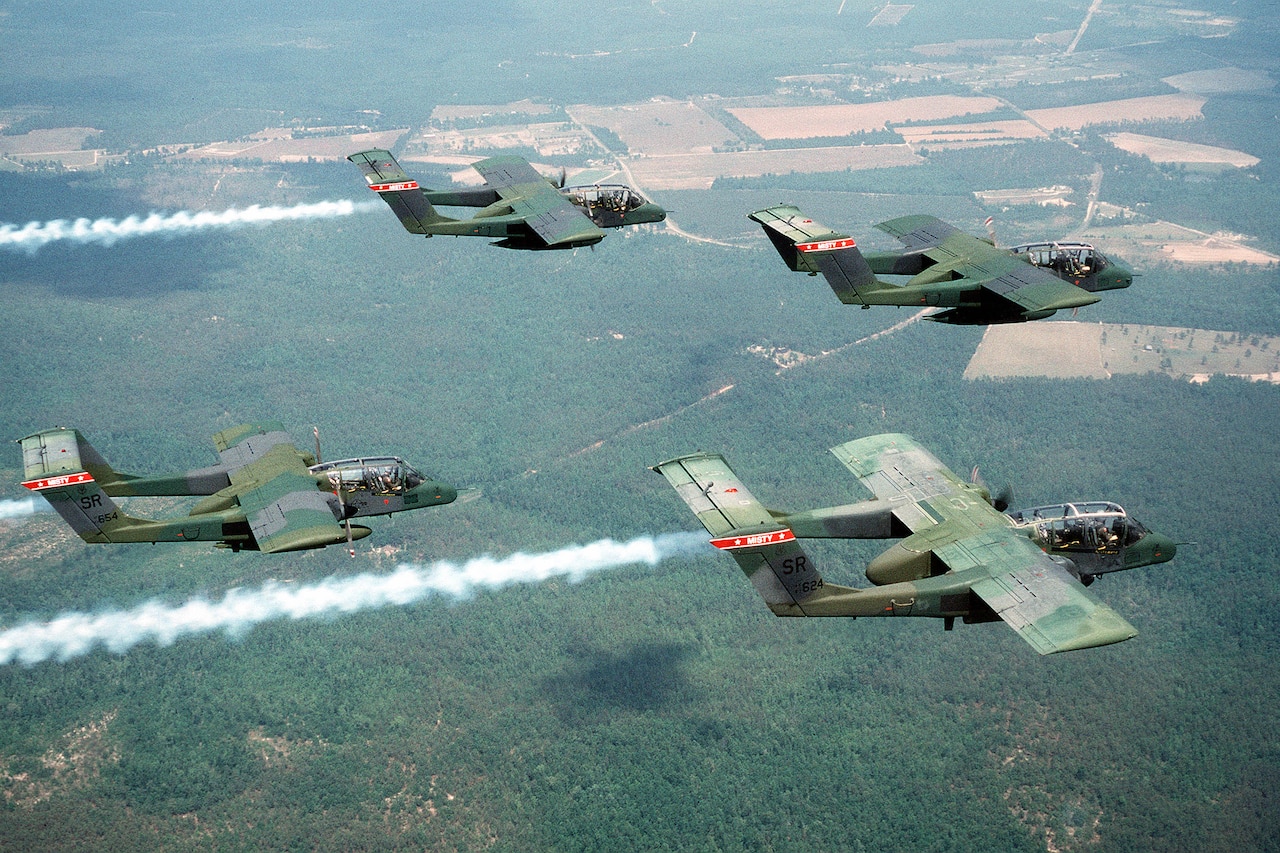 Four camouflage aircraft fly over a dense forest.