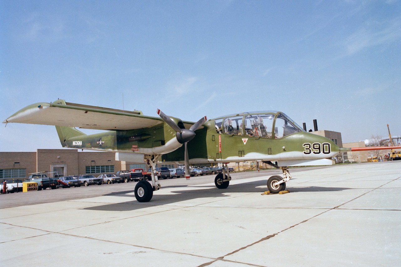 A lightweight propeller aircraft, painted green and gray, is parked on a tarmac. There are several parked cars and a building in the background.