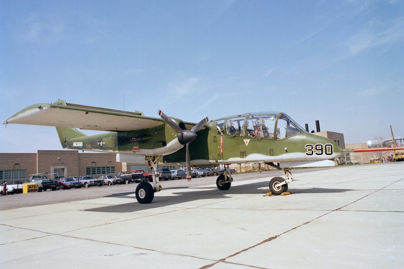 A lightweight propeller aircraft, painted green and gray, is parked on a tarmac. There are several parked cars and a building in the background.