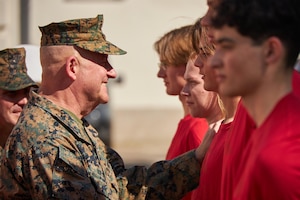 A military officer in a utility uniform stands with his right hand on the shoulder of a young person in a T-shirt. Other service members in similar uniforms are visible in the background.