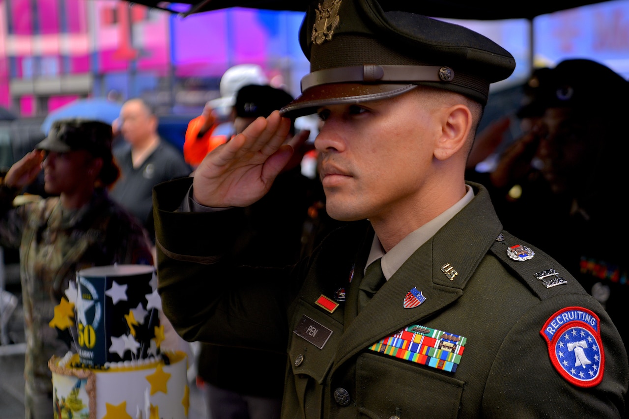 A soldier in a dress uniform with a recruiting patch on the shoulder salutes with his right hand.