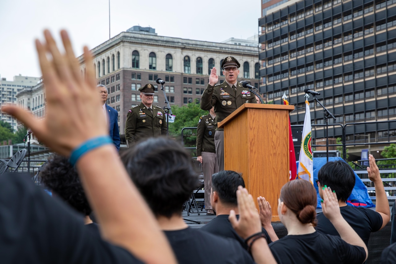 A military officer in a dress uniform stands at a wooden podium, raising his right hand while speaking into a microphone. A crowd of people in black shirts, also with right hands raised, faces him.