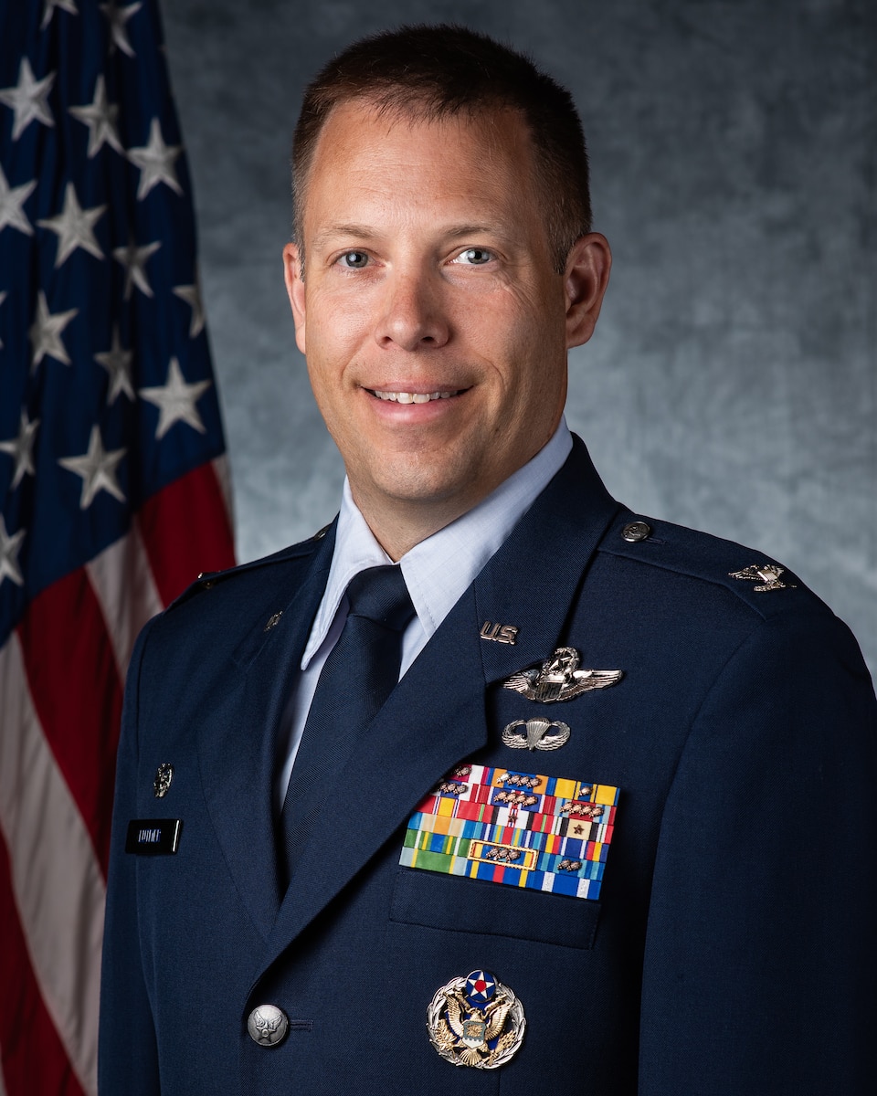 An Air Force fighter pilot in dress blues stands in front of an American flag and a grey background and smiles into the frame.