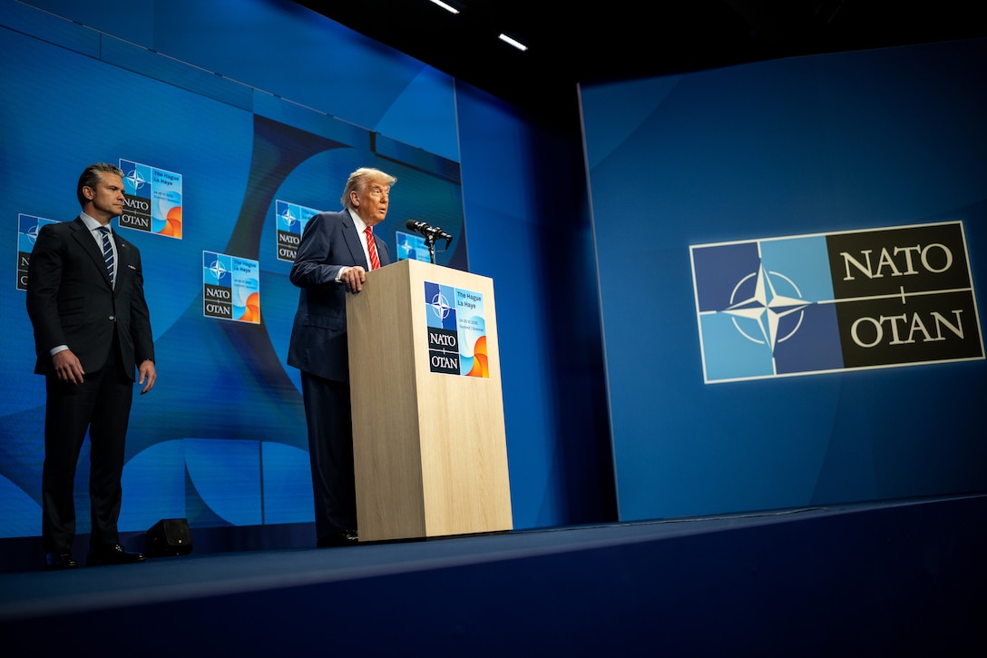 One man in a suit speaks from behind a lectern while another stands behind him. A blue screen with NATO and OTAN is the background.