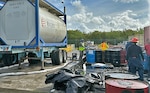 Men use a pump to empty barrels into a large tank in an industrial setting.