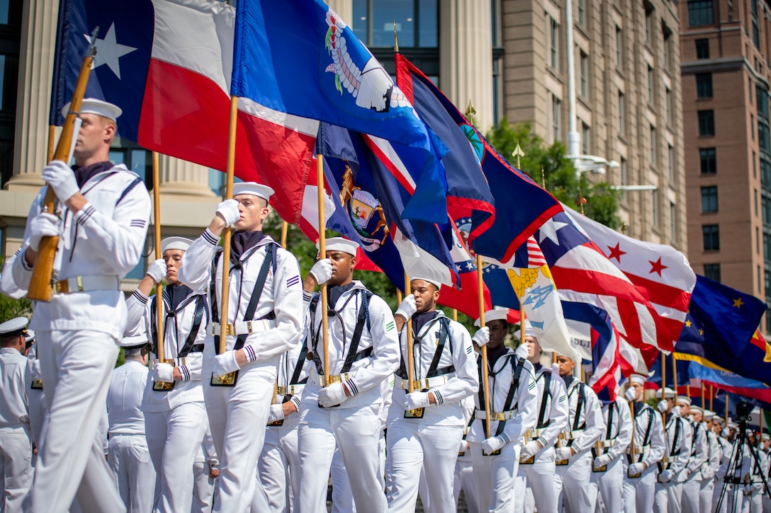 Sailors in uniform hold flags and walk in formation on a street.